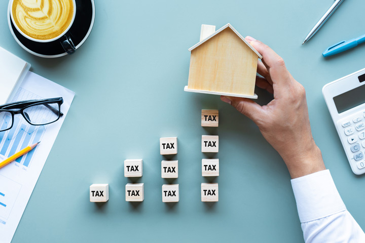 Top down perspective of a desk, man holds a miniature home above an upwards trending graph formation of dice with the words tax inscribed on each